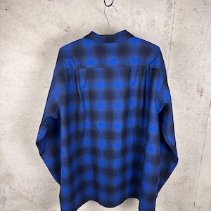 May include: A long-sleeve button-down shirt featuring a bold blue and black ombre plaid pattern. The garment has a classic collar, a straight back yoke, and long sleeves with cuffs, displayed hanging from a metal hook against a neutral wall.