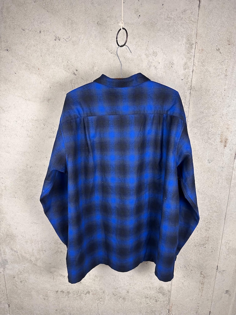 May include: A long-sleeve button-down shirt featuring a bold blue and black ombre plaid pattern. The garment has a classic collar, a straight back yoke, and long sleeves with cuffs, displayed hanging from a metal hook against a neutral wall.