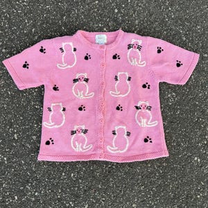 May include: A short-sleeved pink knit cardigan featuring a repeating pattern of embroidered white cats with black whiskers and small black paw prints. The garment has a round neckline, a front button closure, and a ribbed trim along the edges.