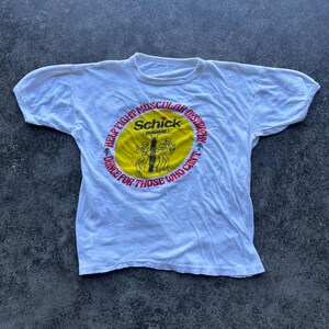 May include: White t-shirt with a crew neck and short sleeves. The shirt features a circular graphic with the text "HELP FIGHT MUSCULAR DYSTROPHY" and "DANCE FOR THOSE WHO CAN'T". The graphic also includes the brand name "Schick".