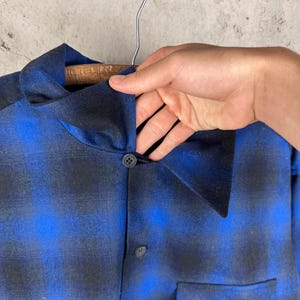 May include: A long-sleeve shirt featuring a blue and black ombre plaid pattern. The shirt has a pointed collar, a front button placket with dark circular buttons, and two large patch pockets on the lower front section of the garment.