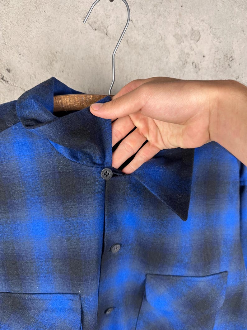 May include: A long-sleeve shirt featuring a blue and black ombre plaid pattern. The shirt has a pointed collar, a front button placket with dark circular buttons, and two large patch pockets on the lower front section of the garment.