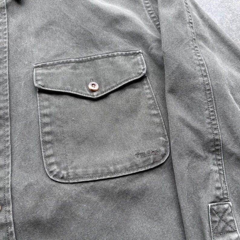 May include: Close-up of a dark green button-down shirt with a single chest pocket. The pocket has a button closure and the brand name "FILSON" is embroidered on the pocket. The shirt is made of a durable fabric.