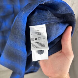 May include: A blue and black plaid shirt featuring a white care label with the text: RN 29685, SHELL: 100% VIRGIN WOOL, LINING: 65% POLYESTER, 35% COTTON, MACHINE WASH WITH LIKE COLORS, COLD, GENTLE CYCLE. NO BLEACH. LINE DRY. STEAM PRESS WITH WARM IRON.