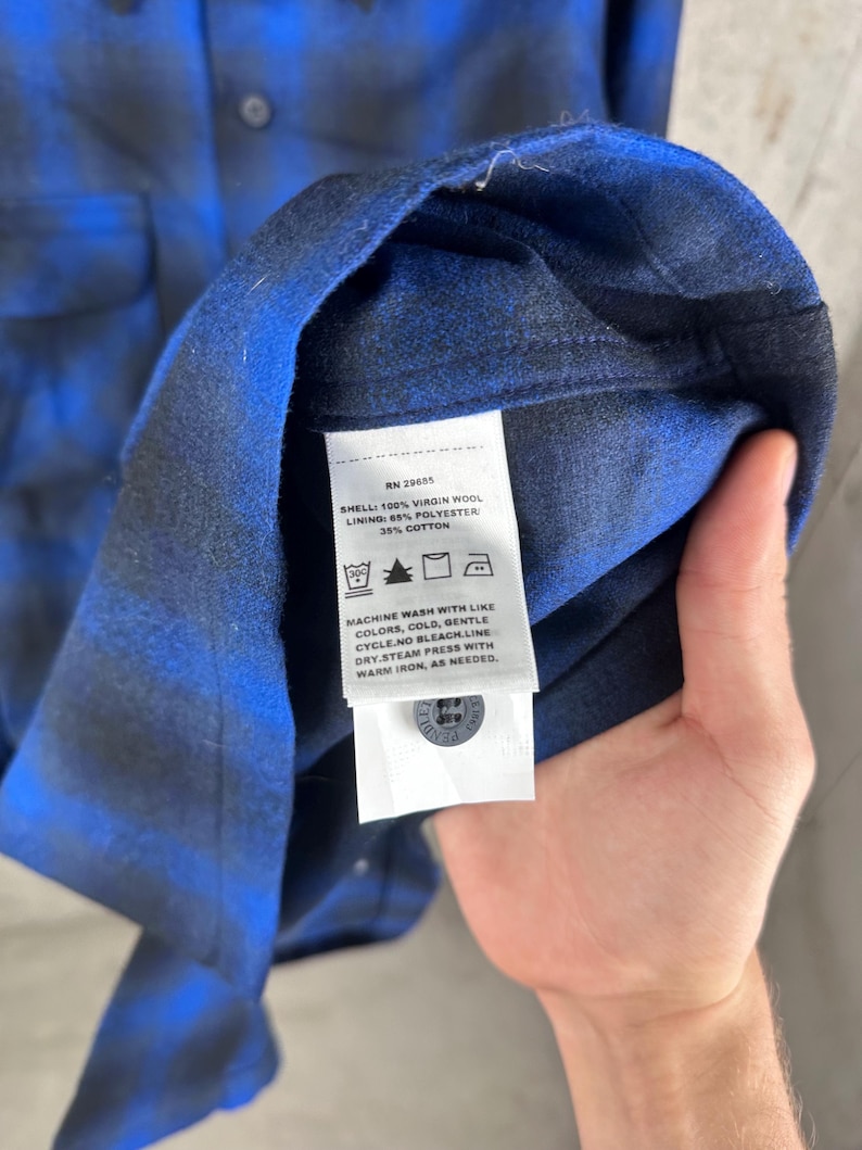 May include: A blue and black plaid shirt featuring a white care label with the text: RN 29685, SHELL: 100% VIRGIN WOOL, LINING: 65% POLYESTER, 35% COTTON, MACHINE WASH WITH LIKE COLORS, COLD, GENTLE CYCLE. NO BLEACH. LINE DRY. STEAM PRESS WITH WARM IRON.
