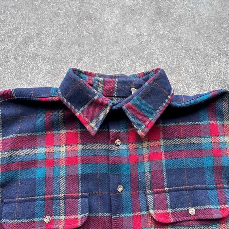 K&ouml;nnte beinhalten: Nahaufnahme eines marineblauen Flanellhemdes mit einem rot, t&uuml;rkis und hellblau karierten Muster. Das Hemd hat eine Knopfleiste, zwei Brusttaschen und einen Kragen. Der Stoff ist weich und warm.
