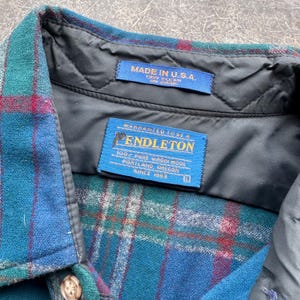 Op de afbeelding: Een close-up van een kraag van een geruit overhemd in teal, blauw en rood. Het overhemd heeft een zwarte gewatteerde voering en twee blauwe labels met de tekst "MADE IN U.S.A." en "PENDLETON 100% PURE VIRGIN WOOL PORTLAND, OREGON SINCE 1863".
