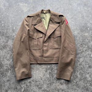 May include: A vintage olive-brown military jacket with a notched collar, four front pockets, and a waist belt. The jacket has a red emblem on the upper left sleeve. The interior lining is a light green color.