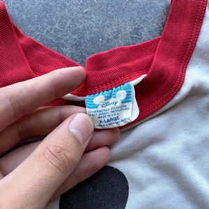 May include: Close-up of a vintage Disney t-shirt. The shirt is off-white with a red neckline and sleeve trim. The tag reads "Disney Character Fashions, Walt Disney Productions, Made in USA, X-Large".