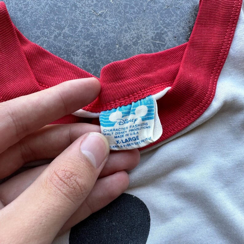 May include: Close-up of a vintage Disney t-shirt. The shirt is off-white with a red neckline and sleeve trim. The tag reads "Disney Character Fashions, Walt Disney Productions, Made in USA, X-Large".