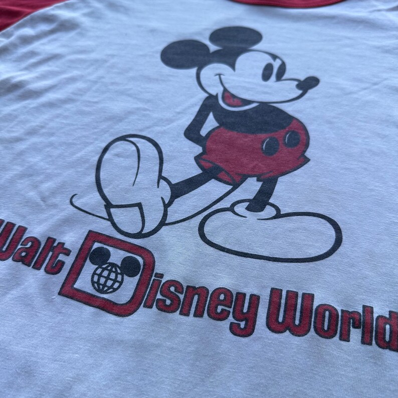 May include: A light gray t-shirt with a red neckline and sleeves. The shirt features a large graphic of Mickey Mouse in red shorts and a black outline. The text "Walt Disney World" is printed in red.
