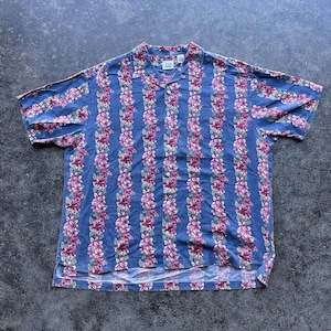 May include: A short-sleeved button-down shirt in a medium blue color featuring vertical stripes of pink and white hibiscus flowers. The shirt has a classic collar, a single chest pocket on the left side, and a white Gap brand label at the neck.