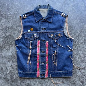 May include: A vintage denim vest in a medium wash, featuring a collared neckline and button-up front. The vest is adorned with decorative pins, pink floral ribbon accents, and dangling leather ties. The vest has a distressed look.