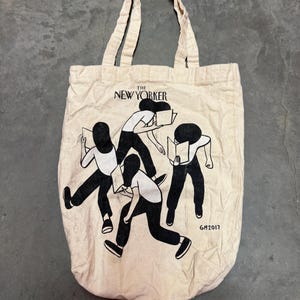 May include: A beige canvas tote bag featuring the text "THE NEW YORKER" above a black and white illustration of figures holding books. The bag has two beige handles and the year "GM2017" printed on the bottom right.
