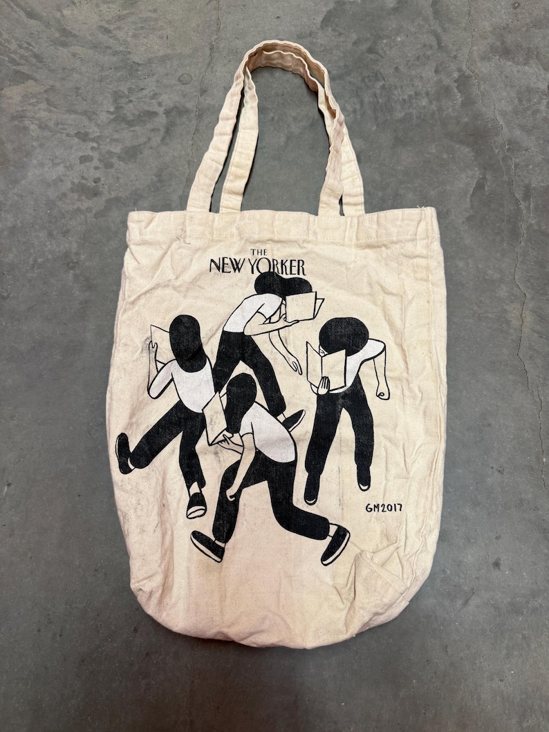 May include: A beige canvas tote bag featuring the text "THE NEW YORKER" above a black and white illustration of figures holding books. The bag has two beige handles and the year "GM2017" printed on the bottom right.