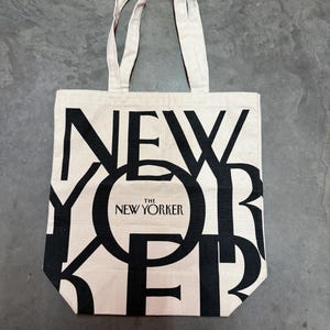 May include: A beige tote bag with black text that reads "NEW YORKER" in a bold, overlapping design. The text "THE NEW YORKER" is centered within the "O" of "YORKER". The bag has two beige handles.