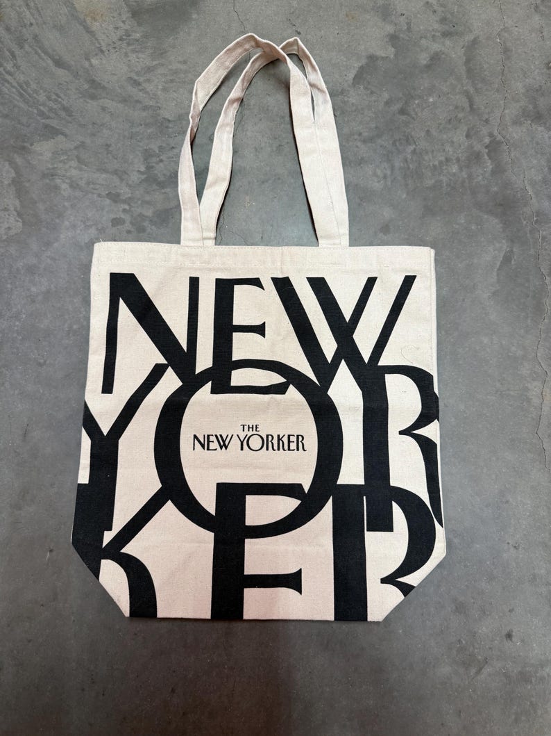 May include: A beige tote bag with black text that reads "NEW YORKER" in a bold, overlapping design. The text "THE NEW YORKER" is centered within the "O" of "YORKER". The bag has two beige handles.