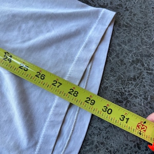 May include: A light-colored t-shirt with a yellow measuring tape laid across it. The tape shows measurements from 24 to 32 inches. The shirt's edge is visible, showing the seam and fabric texture.