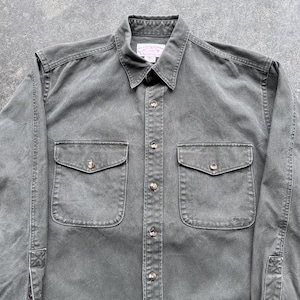 May include: A dark gray button-down shirt with two chest pockets and a collar. The shirt has long sleeves and a classic design. The buttons are a light brown color. The shirt is made of a durable fabric.