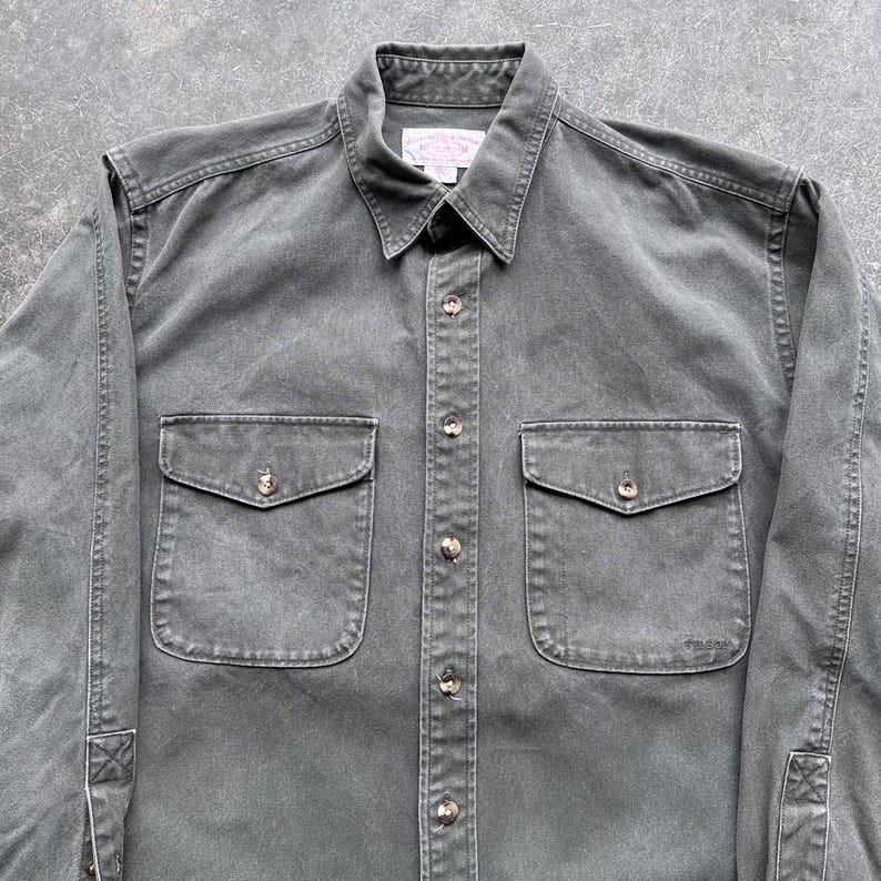 May include: A dark gray button-down shirt with two chest pockets and a collar. The shirt has long sleeves and a classic design. The buttons are a light brown color. The shirt is made of a durable fabric.