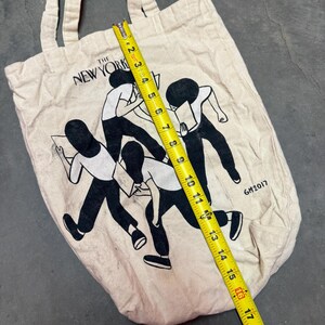 May include: A beige canvas tote bag with the text "THE NEW YORK" and a black and white illustration of figures reading. The bag is approximately 16 inches long. The year "GM2017" is printed on the bag.