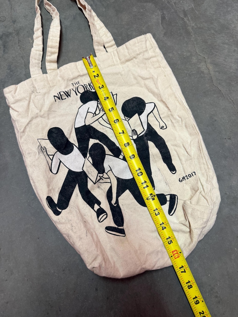 May include: A beige canvas tote bag with the text "THE NEW YORK" and a black and white illustration of figures reading. The bag is approximately 16 inches long. The year "GM2017" is printed on the bag.