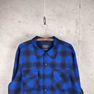 May include: A long-sleeve button-up shirt featuring a blue and black plaid pattern. The shirt has a pointed collar, two chest pockets with flaps, and a dark interior label that reads Pendleton, The Original Board Shirt, size XL.