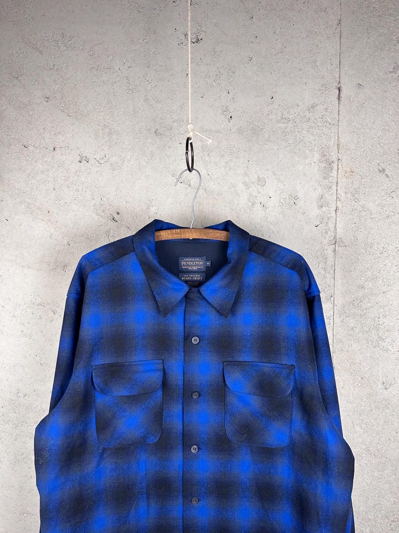 May include: A long-sleeve button-up shirt featuring a blue and black plaid pattern. The shirt has a pointed collar, two chest pockets with flaps, and a dark interior label that reads Pendleton, The Original Board Shirt, size XL.