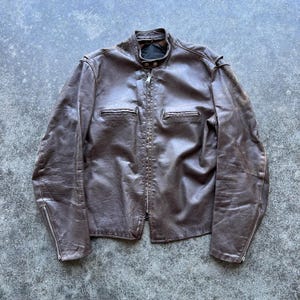 May include: A dark brown leather jacket featuring a stand-up collar with a snap button closure, a full-length front zipper, two horizontal zippered chest pockets, and long sleeves with zippered cuffs. The leather has a visible textured grain.