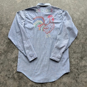 May include: Light blue long-sleeve shirt with a button-down collar. The back features a colorful embroidered rooster design and the word "DECOLORES" in rainbow thread. The shirt has a curved hemline.