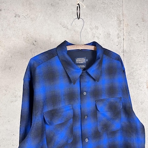 May include: A long-sleeve button-up shirt featuring a blue and black ombre plaid pattern. The shirt has a pointed collar, two front chest pockets with flaps, and a Pendleton brand label on the inside neck. The fabric has a soft, brushed texture.