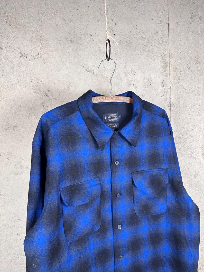 May include: A long-sleeve button-up shirt featuring a blue and black ombre plaid pattern. The shirt has a pointed collar, two front chest pockets with flaps, and a Pendleton brand label on the inside neck. The fabric has a soft, brushed texture.