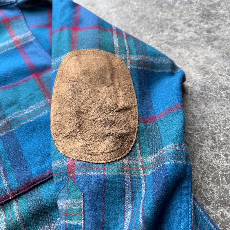 Op de afbeelding: Close-up van een blauw, groen en rood geruit flanellen overhemd met een bruine su&egrave;de ellebooglap. Het overhemd heeft een button-down kraag en een zak. De stof heeft een zachte, getextureerde uitstraling.