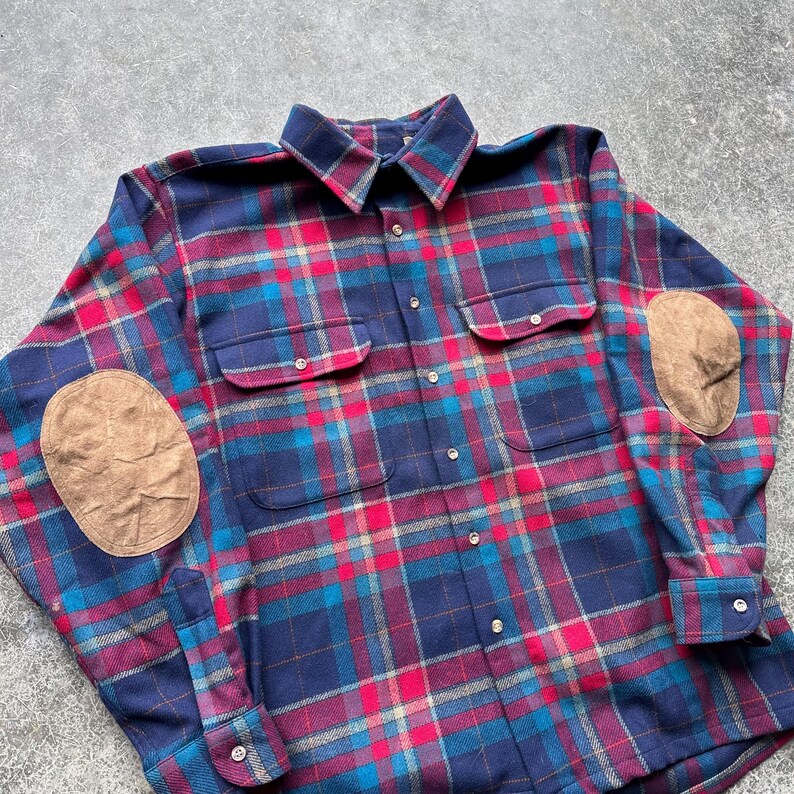 K&ouml;nnte beinhalten: Ein lang&auml;rmliges kariertes Hemd im klassischen Stil. Das Hemd hat ein marineblaues, rotes und t&uuml;rkisfarbenes Karomuster mit braunen Wildleder-Ellbogenpatches. Es hat zwei Brusttaschen und eine Knopfleiste.