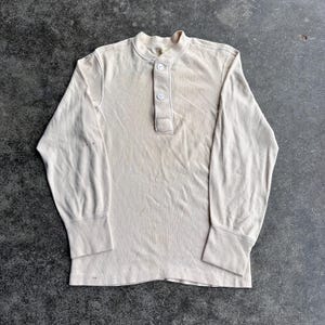 May include: A long-sleeved, off-white shirt with a Henley neckline and three buttons. The shirt has ribbed cuffs and a slightly worn appearance, suggesting vintage or pre-owned condition. The fabric appears to be a soft, knit material.