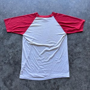 May include: A white and red baseball style t-shirt. The shirt has a white body with red sleeves and a red neckline. The shirt is made of a soft, lightweight material.