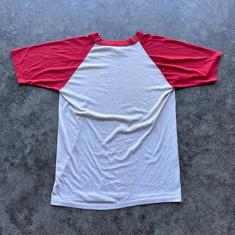 May include: A white and red baseball style t-shirt. The shirt has a white body with red sleeves and a red neckline. The shirt is made of a soft, lightweight material.