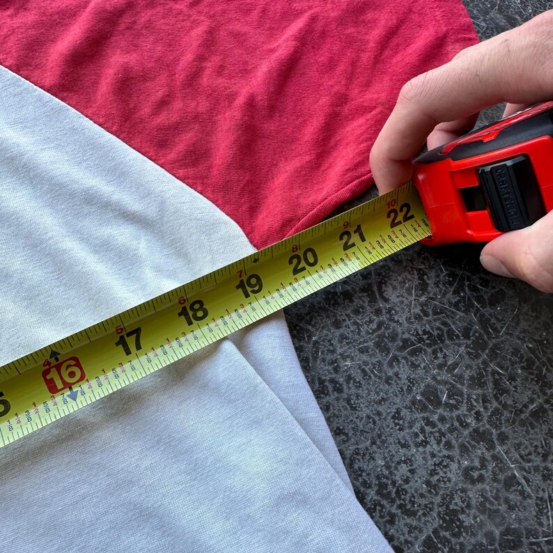 May include: A white and red raglan shirt is measured with a yellow tape measure. The tape measure shows a measurement of approximately 16 to 22 inches. The shirt is made of a soft material.