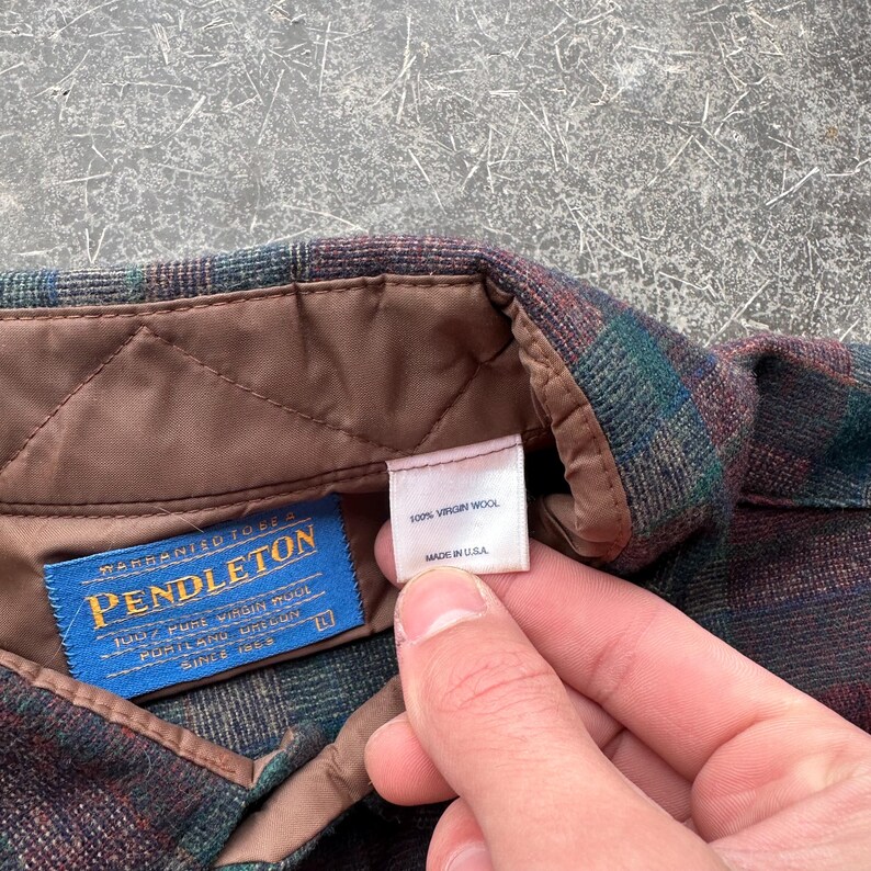 May include: Close-up of a Pendleton wool shirt label. The label reads "PENDLETON" in blue with gold lettering. The shirt is a plaid pattern in shades of green, brown, and burgundy. The label also states "100% Virgin Wool" and "Made in USA."