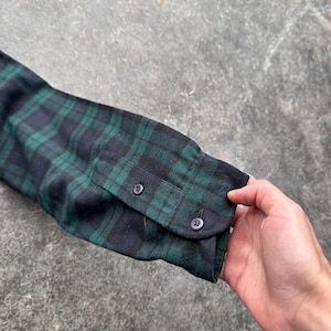 May include: Close-up of a long-sleeved shirt sleeve in a green and navy blue plaid pattern. The cuff is buttoned with two small, dark buttons. The fabric appears to be a soft flannel.