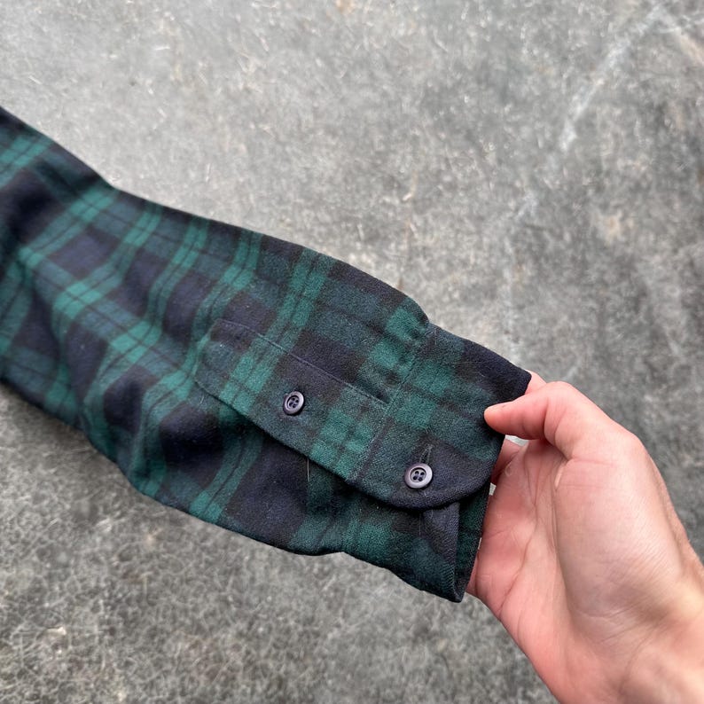 May include: Close-up of a long-sleeved shirt sleeve in a green and navy blue plaid pattern. The cuff is buttoned with two small, dark buttons. The fabric appears to be a soft flannel.