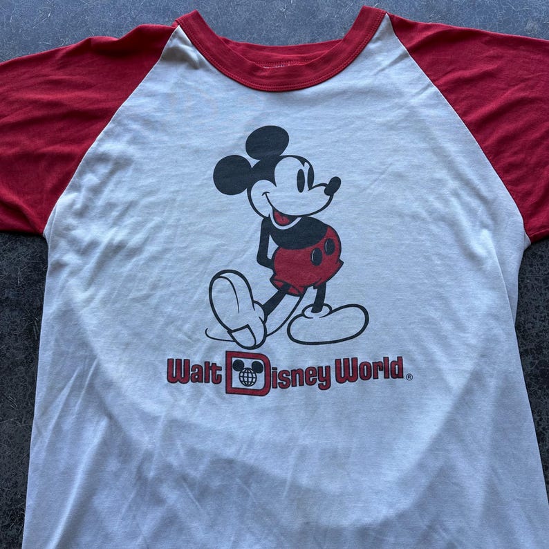 May include: A white and red raglan t-shirt featuring a graphic of Mickey Mouse in black, red, and white. The text "Walt Disney World" is printed in red below the character.