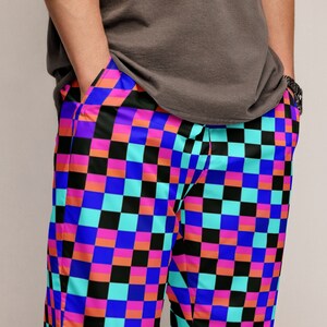 May include: Vibrant, checkered pattern pants in a bold color scheme of blue, pink, orange, black, and turquoise. The pants have pockets and a relaxed fit. The person is wearing a gray t-shirt.