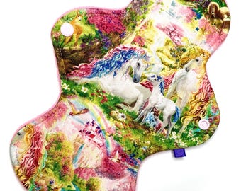 May include: A reusable cloth menstrual pad with a colorful unicorn and rainbow design. The pad features a pink trim, snap closures, and a purple tag. The artwork includes a castle, birds, and a rainbow.