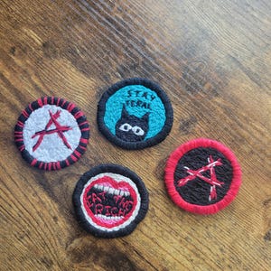 May include: Four embroidered patches with black, red, and white thread. One patch features a black cat with the text "Stay feral". Another patch features a red mouth with the text "Eat the rich". The other two patches feature a red and white symbol.