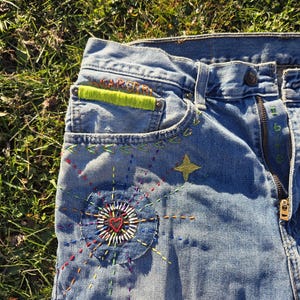 May include: Close-up of blue denim jeans with colorful embroidered details. The jeans feature a patch with a heart design, a star, and the text "WEAR OUT". The stitching is in various colors, adding a unique, handcrafted look.