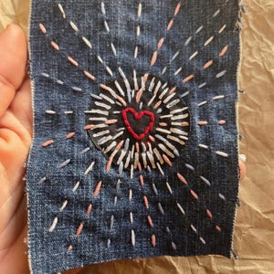 May include: Embroidered denim patch featuring a central heart design. The heart is red, surrounded by a black circle, and radiating white, pink, and blue stitches. The patch is square and has a raw edge.