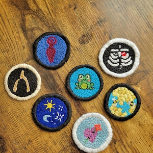May include: Seven embroidered patches with various designs. The patches are circular and feature a variety of colors, including blue, green, yellow, white, and black. The designs include a frog, a beehive, a heart, a moon and stars, a wishbone, a skeleton, and a female figure.
