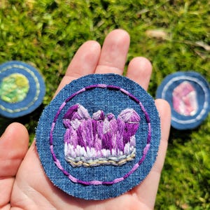 May include: A purple amethyst crystal embroidered on a blue denim circle patch.