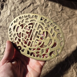 Puede incluir: Un salvamanteles ovalado de latón con un diseño calado. El salvamanteles presenta un patrón repetitivo de formas geométricas. El latón tiene una pátina cálida y envejecida. El salvamanteles se sostiene en una mano.